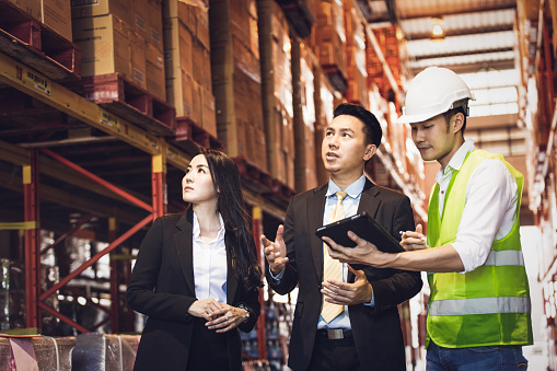 Asian male foreman or manager showing report from digital tablet to business man and woman or CEO in warehouse company. people discuss of logistics shipping, quality control or export import goods.
